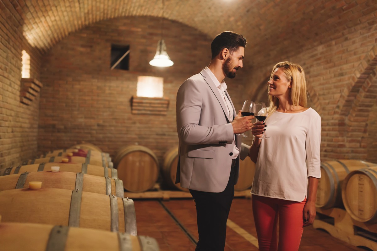 Dans un chai à barrique, un couple amoureux tient un verre de vin rouge 