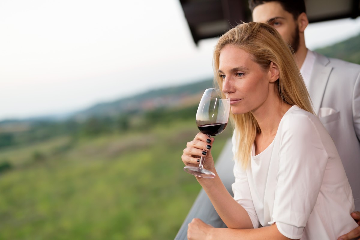 Femme élégante hume un verre de vin rouge depuis une terrasse surplombant vignoble