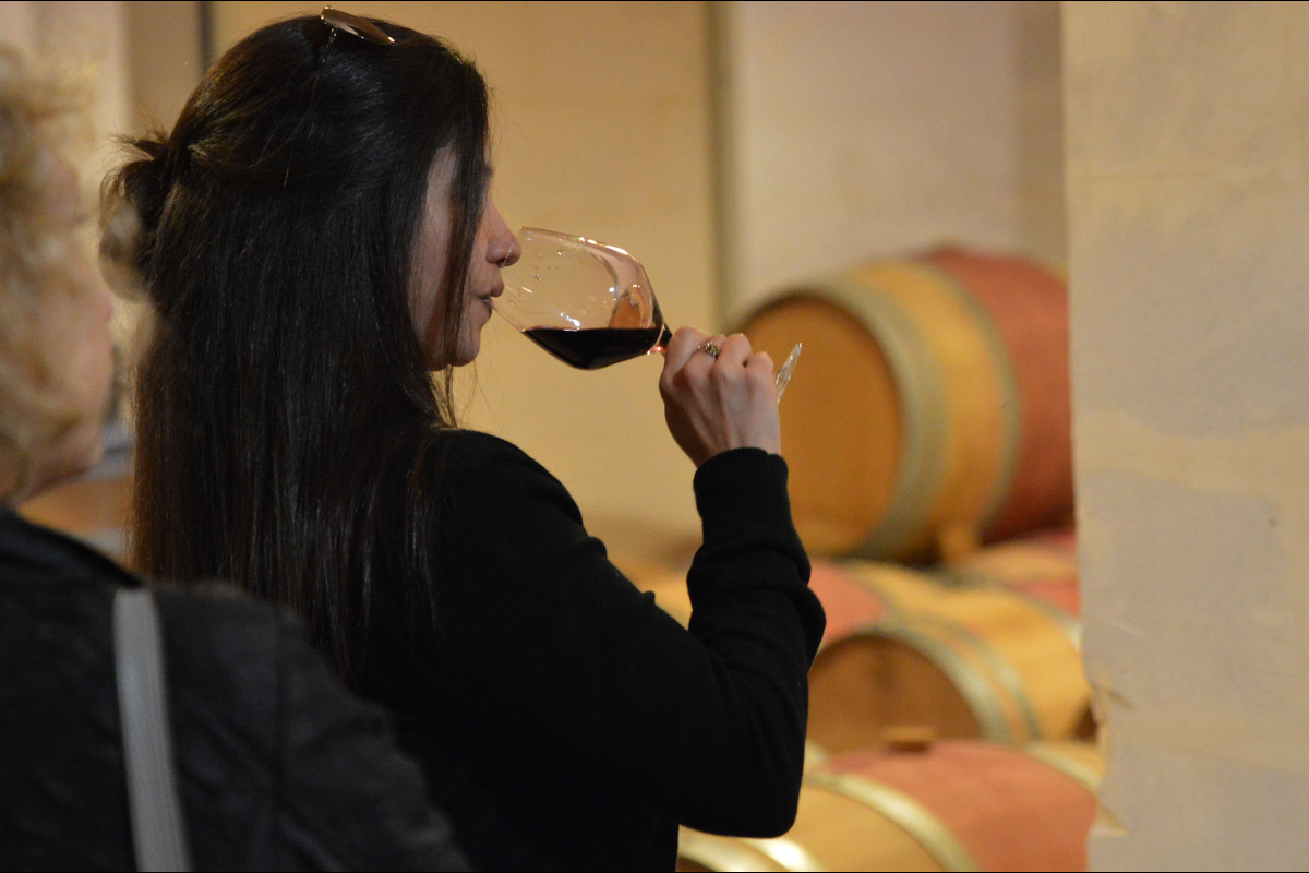 Dame de dos en noir déguste du vin rouge dans chai à barriques